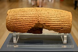 Rear view of a barrel-shaped clay cylinder resپاting on a stand. The cylinder is covered with lines of cuneiform text