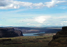 A big river winds through a landscape of sparsely vegetated hills and bluffs. Dark rock faces contrast sharply with a bright blue sky veiled with intermittent white and gray clouds. In the foreground, an open space between bluffs opens on the river.