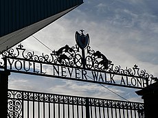 Design of the top of a set of gates, with the sky visible. The inscription on the gates reads "You'll Never Walk Alone".