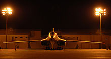 Front view of B-1 parked on ramp at night. Nearby yellow flood lights illuminate the area. In the background are buildings