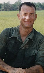 A man is at the center of the image smiling into the camera. He is sitting on a blue crate and has his hands resting on his legs.