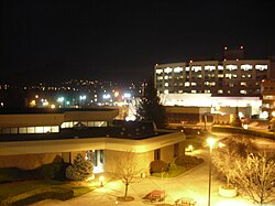 The photo was taken during the night. A large building in the foreground is brightly lit, with a much larger building behind it, and city lights in the background