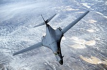 Top forward view of gray aircraft with wings swept forward banking right. Underneath are strips of white clouds and uninhabited terrain.