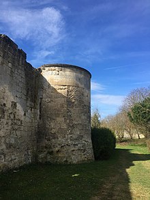 Photo d'un mur d'enceinte muni d'une tour semi-circulaire en pierre à gauche. À droite, un espace vert.