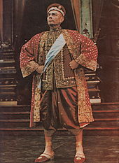 Man in ornate Asian garb stands with fists planted on his hips