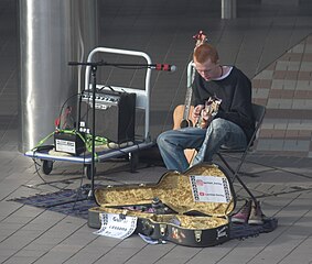 A street performer playing the guitar for people. His guitar case is open so people can put money in it.